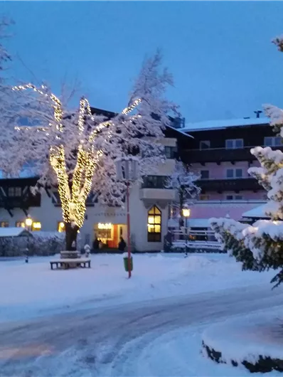 A wintry landscape scene with snow-covered trees and a illuminated building. The twilight creates a cozy atmosphere.