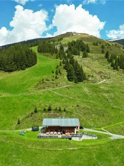 A green mountain landscape with gentle hills and a charming wooden building in the foreground. The sky is blue with some white clouds.