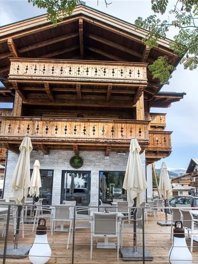 Ein charmantes Holzgebäude mit einem großen Balkon und rustikalem Stil. Vor dem Gebäude befinden sich Tische mit Stühlen und Sonnenschirmen.