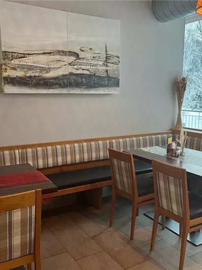 Een gezellig café met houten stoelen en een gestoffeerde bank. Door grote ramen valt licht op het interieur, terwijl buiten een winterlandschap zichtbaar is.