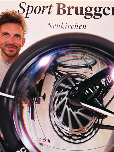 Ein Mann steht neben einem Fahrrad, während er das Rad in die Kamera hält. Im Hintergrund ist das Logo von "Sport Brugger" in Neukirchen sichtbar.