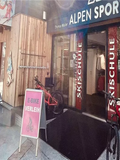Ein Geschäft namens "Alpen Sports" mit Angeboten für Skischule und Fahrräder. Vor dem Laden stehen mehrere Fahrräder und ein Schild.