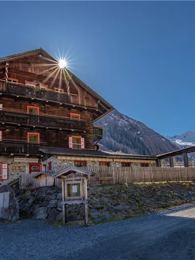 A traditional wooden house in the Alps with a view of snow-covered mountains. The sun shines over the roof of the house and the sky is clear and blue.