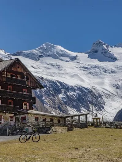 A traditional chalet in the mountains with snow-covered peaks in the background. The surroundings are green and invite outdoor activities.