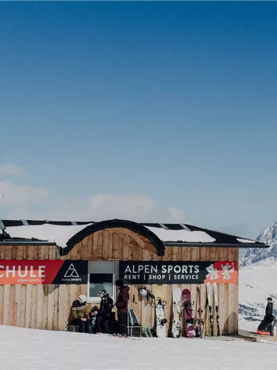 Eine Skischule mit einem Holzgebäude in den Bergen. Schnee bedeckt die Umgebung und der Himmel ist klar und blau.
