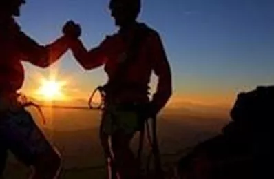 Zwei Bergsteiger feiern den Sonnenuntergang auf einem Gipfel. Sie stehen sich gegenüber und stoßen zusammen, während die Sonne im Hintergrund leuchtet.
