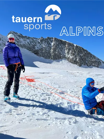 A group of people is practicing with ropes on snow in an alpine environment. In the background, majestic mountains and a clear blue sky can be seen.