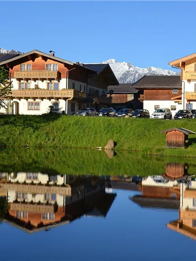 Eine malerische alpine Landschaft mit zwei schönen Häusern am Ufer eines ruhigen Sees. Im Hintergrund sind schneebedeckte Berge und ein klarer blauer Himmel sichtbar.