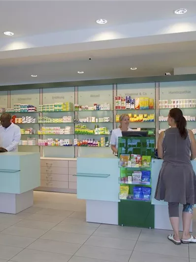 Eine moderne Apotheke mit Regalen voller Produkte. Kunden stehen an derTheke, während Mitarbeiter sie bedienen.