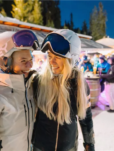 Zwei lächelnde Personen in Skibekleidung stehen sich gegenüber. Im Hintergrund ist eine belebte winterliche Atmosphäre mit Skifahrern und beleuchteten Hütten zu sehen.