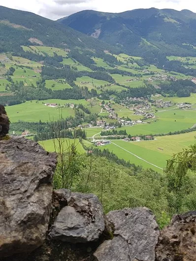 Eine beeindruckende Aussicht auf ein grünes Tal mit kleinen Dörfern und Bergen im Hintergrund. Der Blick ist von Steinen im Vordergrund gerahmt.