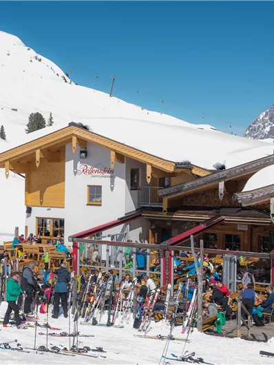 Een gezellig berghuis in de sneeuw met veel skiërs. Op de achtergrond zijn bedekte bergen en een stralend blauwe lucht te zien.