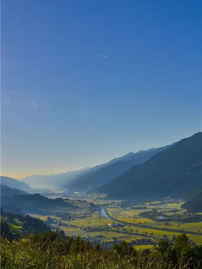 A picturesque landscape with gentle hills and a clear blue sky. In the valley, a river flows, surrounded by meadows and trees.