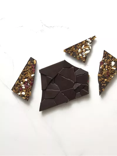Een kleine selectie van chocoladestukjes op een witte marmeren tafel. De chocolade is gedeeltelijk gebroken en wordt omringd door decoratieve, glinsterende stukjes.