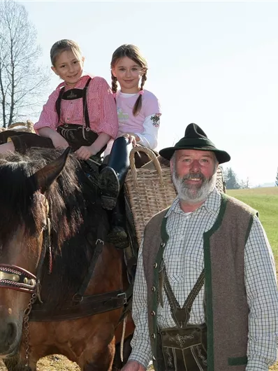 Een man staat naast een paard waarop twee meisjes zitten. De omgeving is landelijk met een heldere lucht en groene weilanden.