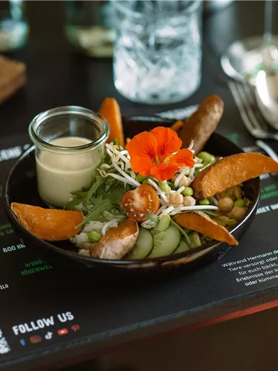 A delicious dish in a bowl that contains vegetables, a blossom, and a small glass with sauce. The table is decorated with a menu and cutlery.