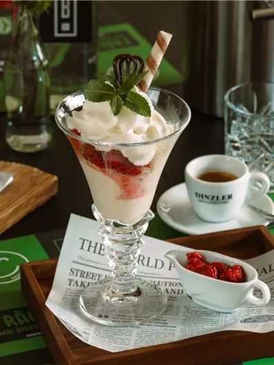 A delicious dessert in a glass with cream, strawberries, and a wafer. There is also a small cup of coffee and a portion of strawberries.