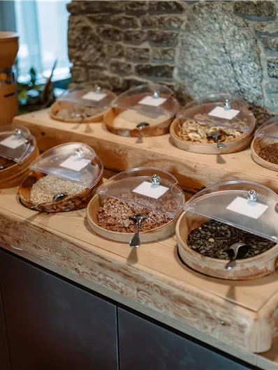 A selection of spices and herbs in glass boxes on a wooden table. In the background, a stone wall is visible.