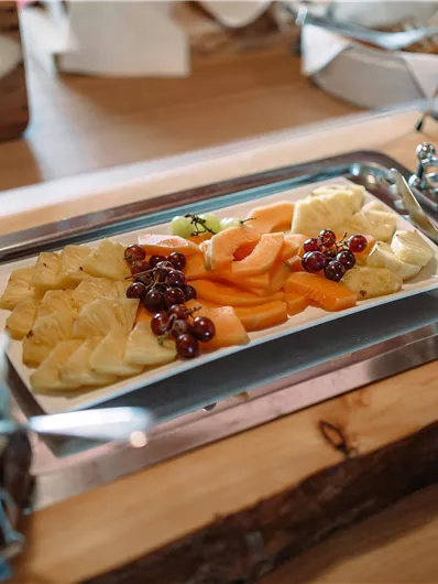 A freshly prepared fruit plate with various fruits such as pineapple, melon, and grapes. In the background, jars of jam are visible.