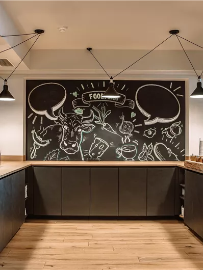 A modern kitchen design with wooden surfaces and a large black chalkboard. The chalkboard displays creative illustrations on the theme of food.