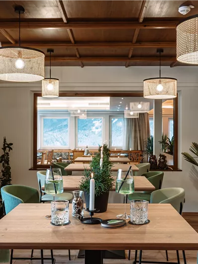 A modern restaurant with wooden furniture and green chairs. Decorative lamps hang on the walls and the room has large windows with a view.