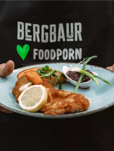 A man is holding a plate with crispy fish, shrimp, and a slice of lemon. In the background, green plants are visible.