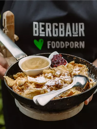A person is holding a pan with freshly prepared food. This includes sauce and a spoon, while green plants are visible in the background.