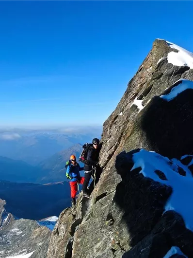 Zwei Bergsteiger klettern auf einen steilen Felsen in den Bergen. Im Hintergrund sind schneebedeckte Gipfel und ein klarer blauer Himmel zu sehen.