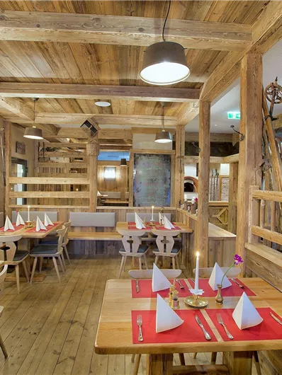 Ein gemütliches Restaurant mit Holzmöbeln und warmem Licht. Die Tische sind mit roten Tischdecken und weißen Servietten dekoriert.