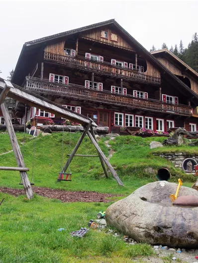 Een rustiek houten huis omringd door groene weiden en bomen. Op de voorgrond bevindt zich een speeltuin met een schommel en speeltoestellen.
