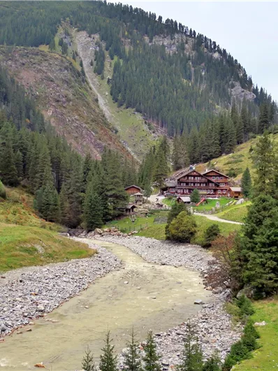 Een schilderachtig berglandschap met bomen en stromend water. Op de achtergrond is een rustiek gebouw zichtbaar.
