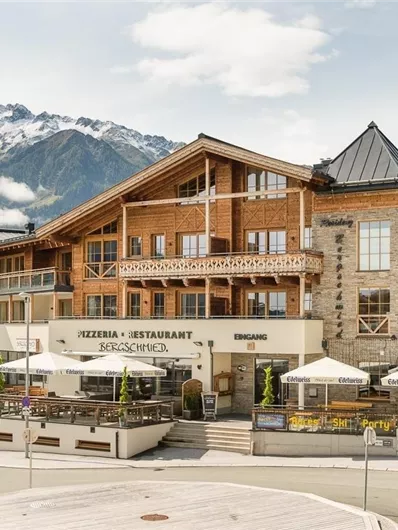Ein traditionelles Bergrestaurant mit einer großen Terrasse und Sonnenschirmen. Im Hintergrund sind majestätische Berge und ein klarer Himmel zu sehen.