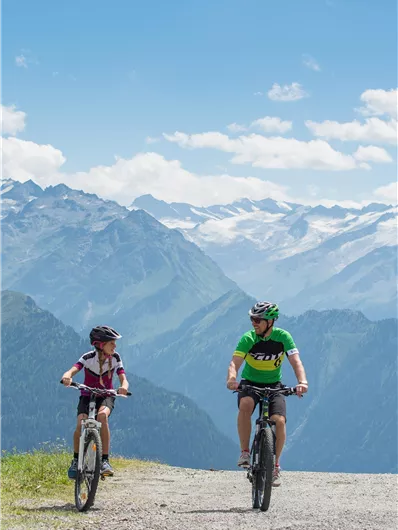 Ein Vater und sein Kind fahren mit Fahrrädern auf einem Schotterweg in den Bergen. Im Hintergrund sind majestätische Berge und ein klarer blauer Himmel zu sehen.