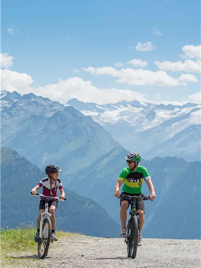 Ein Vater und sein Kind fahren mit dem Fahrrad auf einem Bergweg. Im Hintergrund sind beeindruckende Berge und ein blauer Himmel zu sehen.