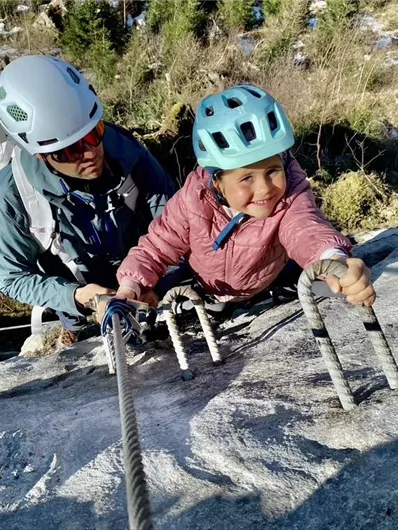 Ein Kletterer sichert einen anderen Kletterer an einer felsigen Wand. Die Szene zeigt einen klaren blauen Himmel und eine beeindruckende Berglandschaft.