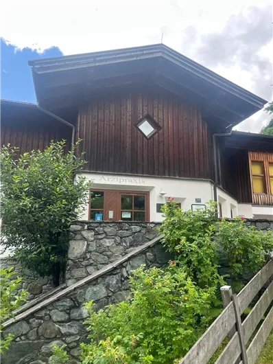 Ein gemütliches Holzhaus mit einem Steinmauer und grünen Büschen. Der Himmel ist teilweise bewölkt.