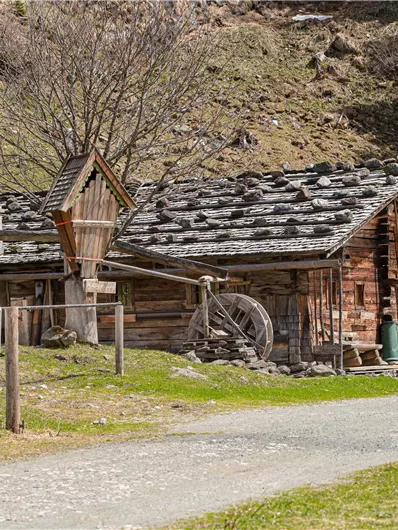 Een rustiek houten huis met een watermolen en een groene tuin. Het landschap wordt gekenmerkt door zachte heuvels.