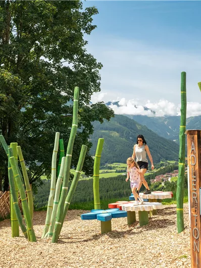 Ein schöner Spielplatz in der Natur mit bunten Elementen und einem herrlichen Aussichtspunkt. Im Hintergrund sind grüne Berge und ein blauer Himmel zu sehen.
