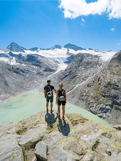 Eine beeindruckende Berglandschaft mit hohen Gipfeln und einem türkisfarbenen See. Zwei Wanderer stehen auf einem Felsen und genießen die Aussicht.