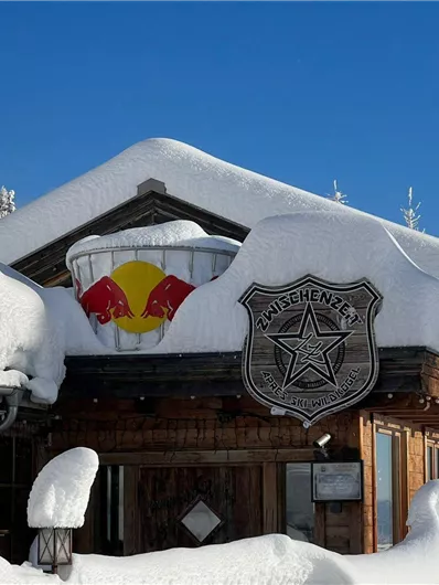 Eine verschneite Berghütte mit einem roten Bull-Logo an der Wand. Der klare blaue Himmel bietet einen schönen Kontrast zur weißen Schneedecke.