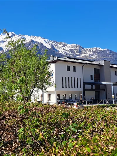 Ein modernes Gebäude vor einer beeindruckenden Bergkulisse. Die Landschaft ist grün und sonnig, mit schneebedeckten Gipfeln im Hintergrund.