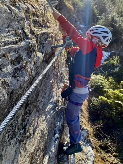 Ein Kletterer sichert sich an einem Felsen mit einem Seil. Im Hintergrund sind Bäume und die Natur zu sehen.