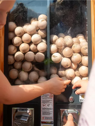 Ein Kind benutzt einen Kaugummiautomaten voller Holzspielbälle. Die Szene zeigt Freude und Neugier im Freien.