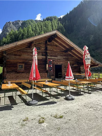 Eine gemütliche Almhütte umgeben von Bergen. Vor der Hütte stehen Holzbänke und Sonnenschirme.