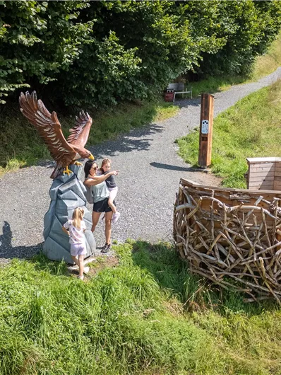 Ein Weg führt durch eine grüne Landschaft. Zwei Kinder stehen neben einer großen Statue eines Adlers und einer Strukturen aus Zweigen.