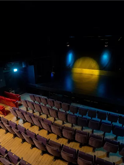 Ein leerer Theatersaal mit rotem und lila Sitzplätzen. Die Bühne ist schwach beleuchtet und hat ein gelbes Licht in der Mitte.