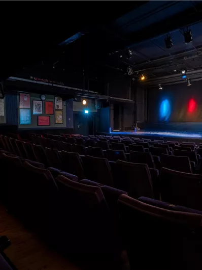 Ein leerer Theatersaal mit roten und blauen Lichtern auf der Bühne. Die Sitze sind ordentlich in Reihen angeordnet.