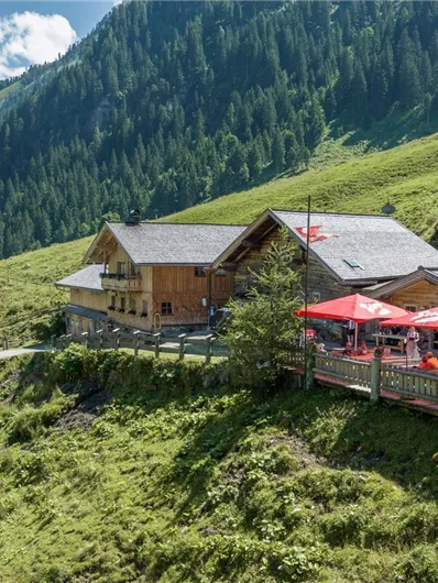 Een Alpenhut in een groene berglandschap. Langs de hut staan rode parasols en de lucht is helder en blauw.