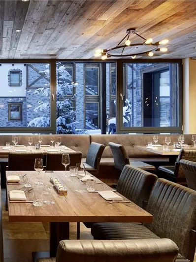 A modern restaurant with wooden elements and large windows. The tables are elegantly set and offer a view of a snowy outdoor area.