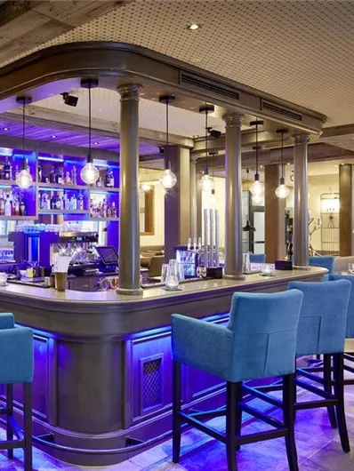 A modern bar with stylish blue chairs and soft lighting. In the background, shelves with various drinks can be seen.